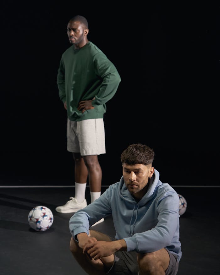 Two young men in casual sportswear with footballs, reflecting in studio setting.